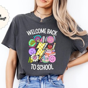 May include: A charcoal grey t-shirt with a graphic print of a lightning bolt and school supplies. The text "Welcome Back To School" is printed in white on the shirt.