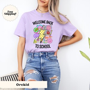 May include: A purple t-shirt with the text "Welcome Back To School" and a graphic of a lightning bolt, a globe, a book, and other school-related items.
