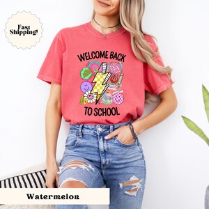 May include: A red t-shirt with a graphic design that says "Welcome Back To School" with a lightning bolt and school-themed icons in pink, yellow, blue, and green.