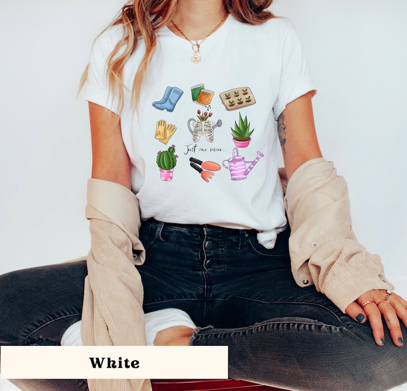 Pu&ograve; includere: Maglietta bianca con una grafica a tema giardinaggio che presenta un annaffiatoio, guanti da giardinaggio, un cactus e il testo "Just one more..."
