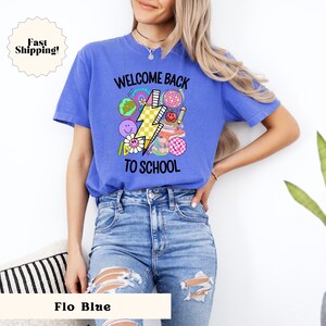 May include: A blue t-shirt with the text "Welcome Back To School" and a graphic of a lightning bolt and school-themed icons. The model is wearing blue jeans.