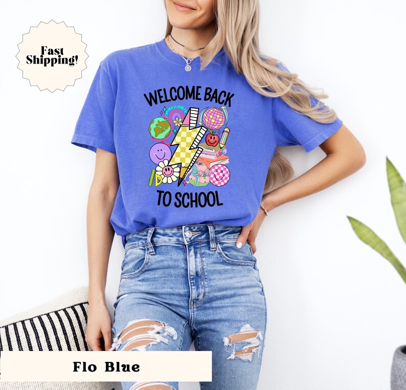 May include: A blue t-shirt with the text "Welcome Back To School" and a graphic of a lightning bolt and school-themed icons. The model is wearing blue jeans.