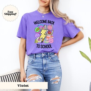 May include: A purple t-shirt with the text "Welcome Back To School" and a graphic of a lightning bolt with school-themed icons. The model is wearing blue jeans.