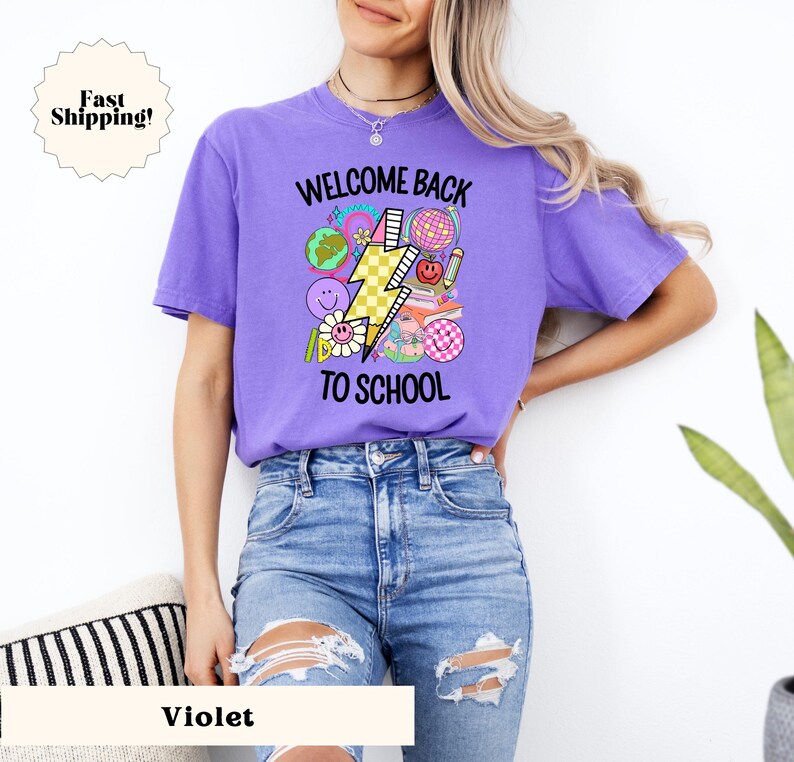 May include: A purple t-shirt with the text "Welcome Back To School" and a graphic of a lightning bolt with school-themed icons. The model is wearing blue jeans.