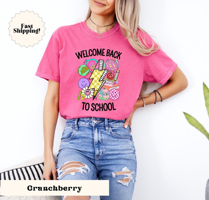 May include: Pink t-shirt with a graphic design of a lightning bolt and school supplies. The text "Welcome Back To School" is printed on the shirt.