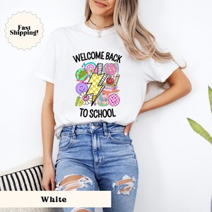 May include: White t-shirt with a graphic design that says "Welcome Back To School" with a lightning bolt and school-themed icons.