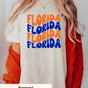May include: A white t-shirt with the word "FLORIDA" repeated four times in a retro, colorful font. The text is in blue and orange. The shirt is on a model wearing a red cardigan.
