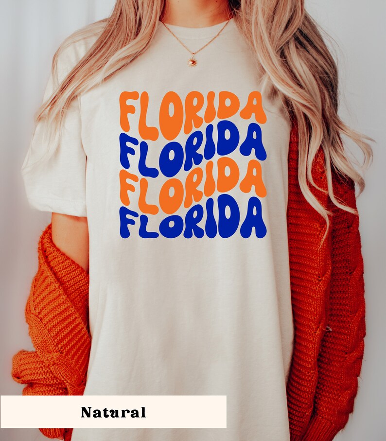 May include: A white t-shirt with the word "FLORIDA" repeated four times in a retro, colorful font. The text is in blue and orange. The shirt is on a model wearing a red cardigan.