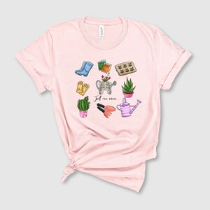 Pu&ograve; includere: Maglietta rosa con un design grafico di attrezzi da giardinaggio e piante. Il testo "Just one more..." &egrave; stampato sulla maglietta.