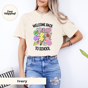 May include: A white t-shirt with a colorful graphic design featuring a lightning bolt, school supplies, and the text "Welcome Back To School".
