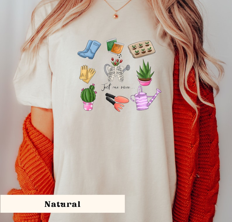 Pu&ograve; includere: Una maglietta bianca con un design grafico di attrezzi da giardinaggio e piante. Il testo "Just one more..." &egrave; stampato sulla maglietta.