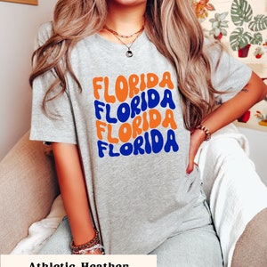 May include: A gray t-shirt with the word "FLORIDA" repeated four times in a retro, wavy font. The text is in orange and blue. The shirt is labeled "Athletic Heather".