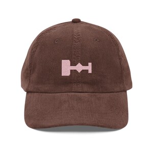 May include: A brown corduroy baseball cap with a pink embroidered symbol on the front. The symbol is a stylized representation of a key.
