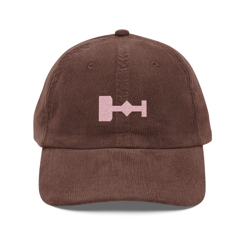 May include: A brown corduroy baseball cap with a pink embroidered symbol on the front. The symbol is a stylized representation of a key.