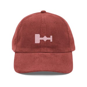 May include: A burgundy corduroy baseball cap with a pink embroidered symbol of a key with a diamond shape in the middle.