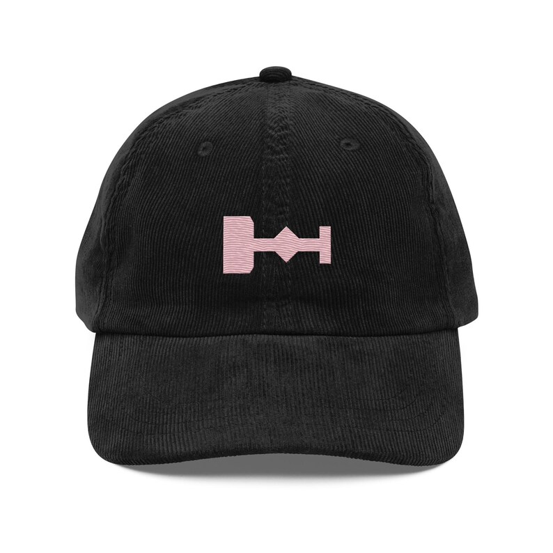 May include: A black corduroy baseball cap with a pink embroidered symbol on the front. The symbol is a stylized geometric shape.
