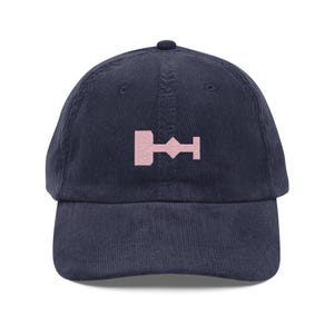 May include: A navy blue corduroy baseball cap with a pink embroidered symbol on the front. The symbol is a stylized geometric shape.
