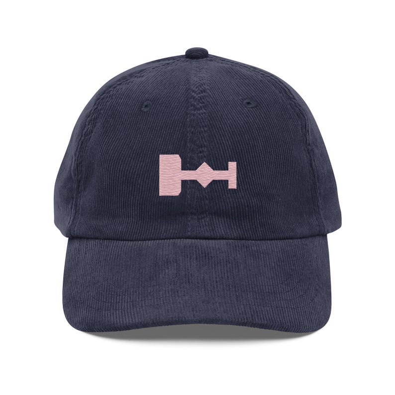 May include: A navy blue corduroy baseball cap with a pink embroidered symbol on the front. The symbol is a stylized geometric shape.