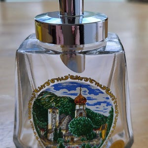 May include: Clear glass cruet with a silver-colored spout and lid. The cruet features a colorful, circular image of a European village with a church. The cruet is sitting on a wooden surface.