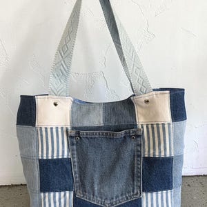 Może przedstawiać: Torba typu tote z patchworku dżinsowego z jasnoniebieskim paskiem z geometrycznym wzorem. Torba ma różne odcienie dżinsu i przednią kieszeń. Torba jest wykonana z dżinsu i ma jasną podszewkę.