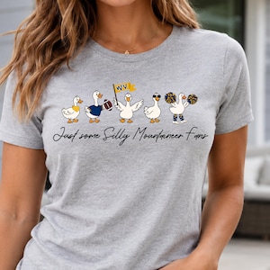 May include: A heather gray t-shirt featuring a graphic of five white ducks in various poses, including one holding a football, one with a WV flag, and one with pom-poms. Below the ducks, the text reads, Just some Silly Mountaineer Fans.