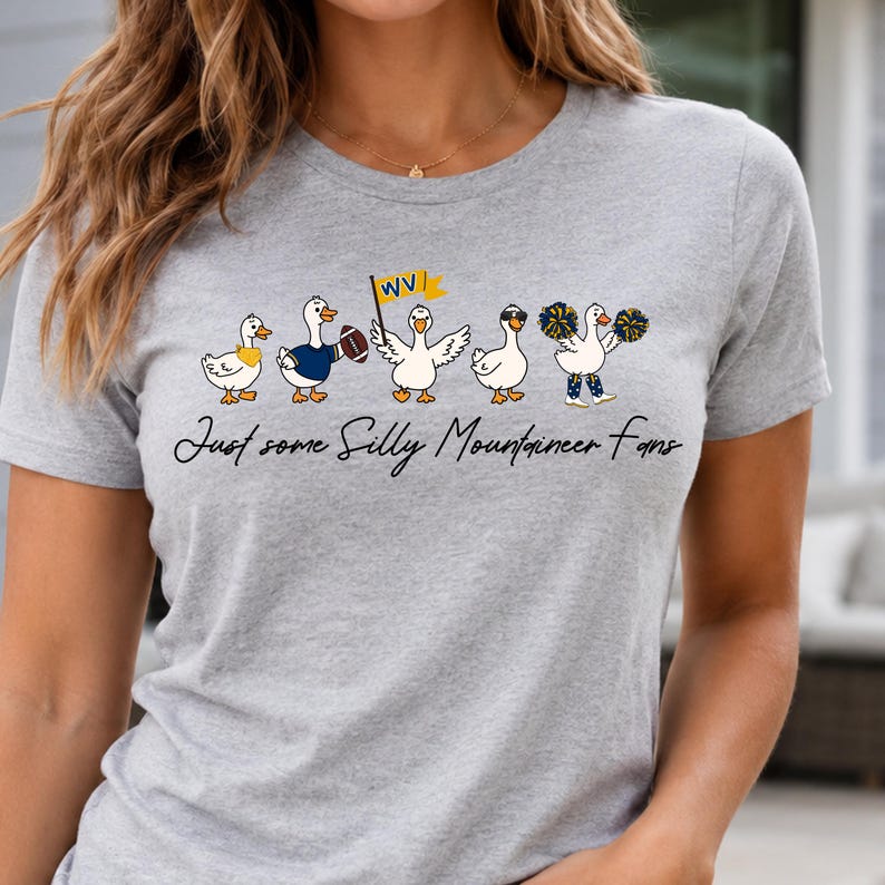 May include: A heather gray t-shirt featuring a graphic of five white ducks in various poses, including one holding a football, one with a WV flag, and one with pom-poms. Below the ducks, the text reads, Just some Silly Mountaineer Fans.