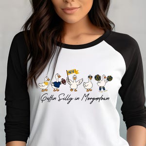 May include: A white raglan shirt with black three-quarter sleeves featuring a graphic of five cartoon ducks. One duck holds a football, another a yellow WV flag, and one wears sunglasses. The text below reads Gettin Silly in Morgantown.