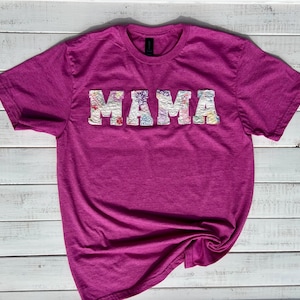 May include: A pink t-shirt with the word "MAMA" spelled out in white fabric with floral patterns.
