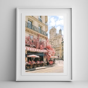 May include: A Parisian cafe with pink flowers blooming outside, a striped awning, and people sitting at tables. The cafe is located on a street with a church in the background.