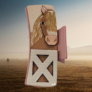 May include: A decorative hair clip featuring a horse design. The clip has a light pink frame and a wooden cutout of a horse's head with a tan mane and a pink nose. The bottom section has a white background with a geometric design.