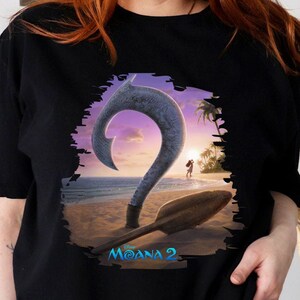 Puede incluir: Camiseta negra con un estampado gr&aacute;fico de un gancho azul y una pala de madera en una playa con una puesta de sol de fondo. El texto "Moana 2" est&aacute; impreso debajo del gr&aacute;fico.