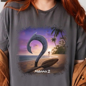 May include: A charcoal gray t-shirt with a graphic print of a cartoon hook and a sunset scene. The text "Moana 2" is printed below the graphic.