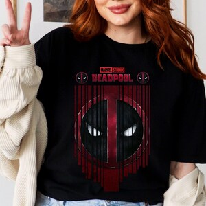 Op de afbeelding: Zwart T-shirt met een rood en zwart Deadpool-logo en de tekst "MARVEL STUDIOS DEADPOOL" gedrukt op de voorkant.