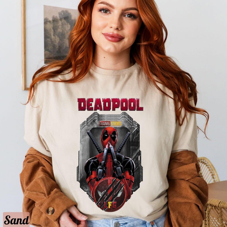 May include: A beige t-shirt with a graphic print of Deadpool from Marvel Studios. The graphic features Deadpool in a red and black suit with his hands up in a fighting pose. The text "DEADPOOL" is printed in red above the graphic.