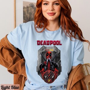 May include: A light blue t-shirt with a graphic of Deadpool from Marvel Studios. The graphic shows Deadpool in a red and black suit with his hands up in a fighting pose. The text "DEADPOOL" is printed in black above the graphic.