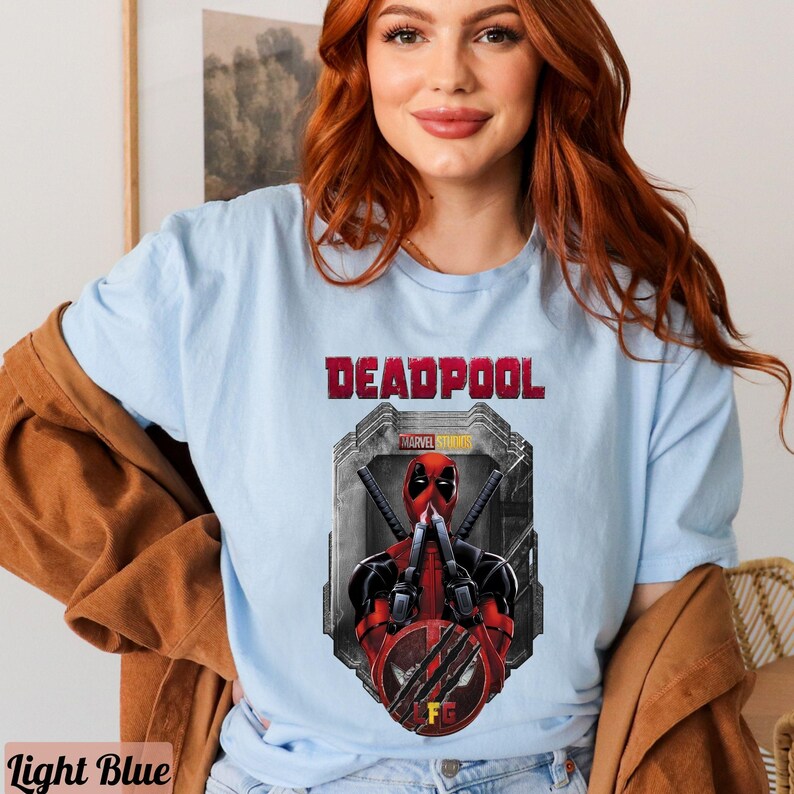 May include: A light blue t-shirt with a graphic of Deadpool from Marvel Studios. The graphic shows Deadpool in a red and black suit with his hands up in a fighting pose. The text "DEADPOOL" is printed in black above the graphic.