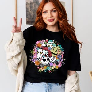 K&ouml;nnte beinhalten: Schwarzes T-Shirt mit einem bunten Grafikdruck von Figuren aus dem Film Nightmare Before Christmas. Die Figuren sind von Blumen umgeben.