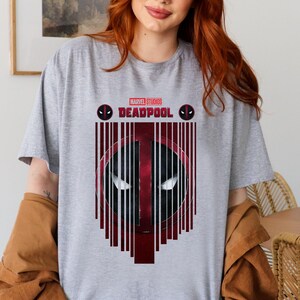 Op de afbeelding: Een grijs T-shirt met een rode en zwarte afbeelding van Deadpool's masker. De afbeelding is omgeven door rode en zwarte verticale lijnen. De tekst "MARVEL STUDIOS DEADPOOL" is in wit boven de afbeelding gedrukt.