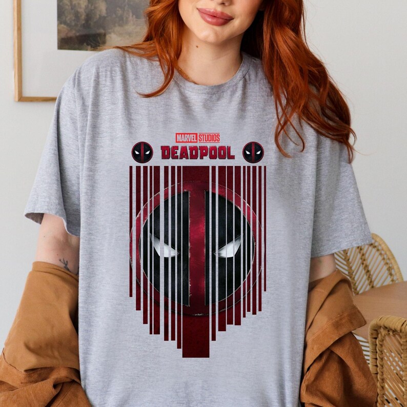 Op de afbeelding: Een grijs T-shirt met een rode en zwarte afbeelding van Deadpool's masker. De afbeelding is omgeven door rode en zwarte verticale lijnen. De tekst "MARVEL STUDIOS DEADPOOL" is in wit boven de afbeelding gedrukt.