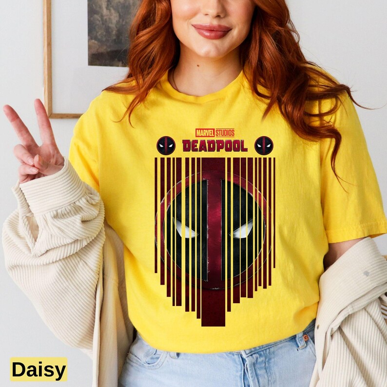 Op de afbeelding: Een geel T-shirt met een zwart-rode afbeelding van Deadpools gezicht met de tekst "MARVEL STUDIOS DEADPOOL" erboven.