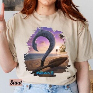 Puede incluir: Una camiseta beige con un estampado gr&aacute;fico de un gancho morado y azul con un fondo de puesta de sol. El texto "Moana 2" est&aacute; impreso en la camiseta.