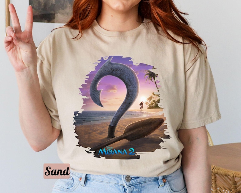 Puede incluir: Una camiseta beige con un estampado gr&aacute;fico de un gancho morado y azul con un fondo de puesta de sol. El texto "Moana 2" est&aacute; impreso en la camiseta.