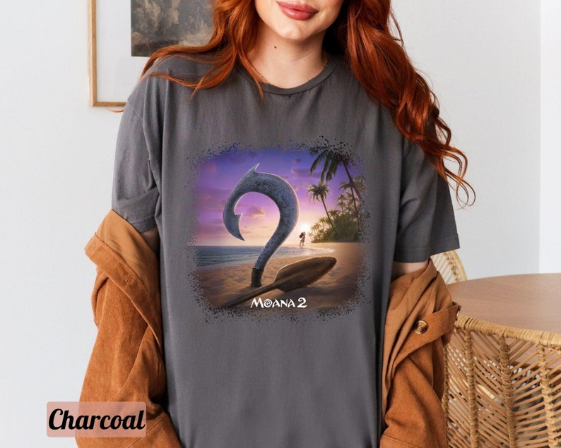 May include: A charcoal gray t-shirt with a graphic print of a cartoon hook and a sunset scene. The text "Moana 2" is printed below the graphic.