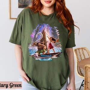 May include: A green t-shirt featuring a graphic print of Moana and Maui sailing on a boat. The graphic is in a cartoon style and has a blue and purple background.