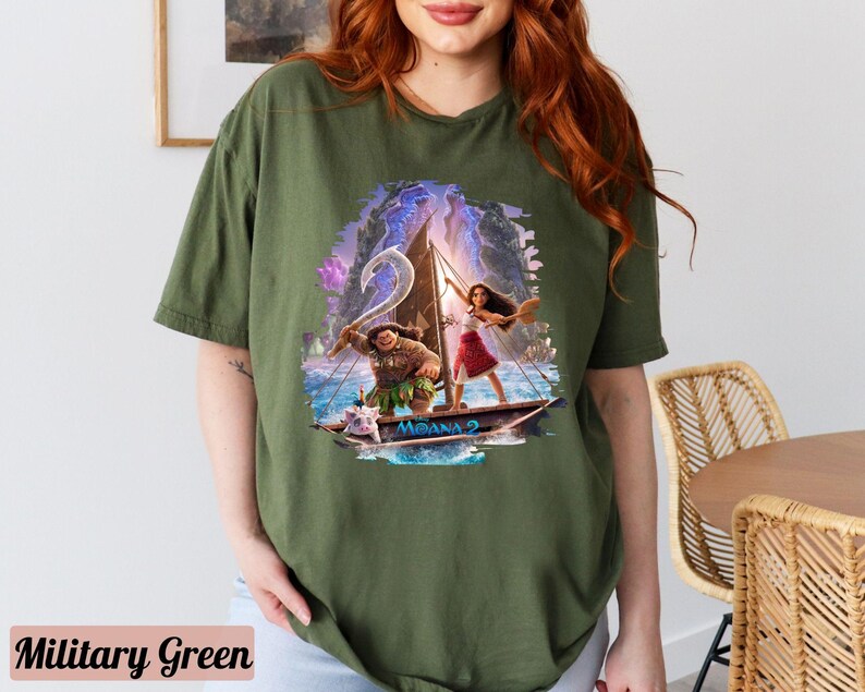 May include: A green t-shirt featuring a graphic print of Moana and Maui sailing on a boat. The graphic is in a cartoon style and has a blue and purple background.