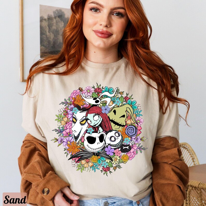 K&ouml;nnte beinhalten: Ein beiges T-Shirt mit einem bunten Grafikdruck von Figuren aus dem Film Nightmare Before Christmas. Die Figuren sind von Blumen und Bl&auml;ttern umgeben.