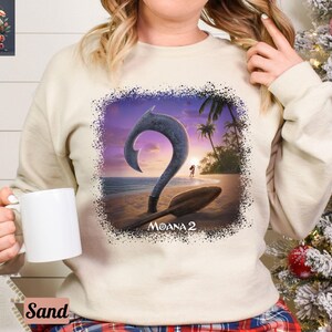 May include: A white sweatshirt with a graphic print of a teal blue ocean scene with a large teal blue hook, a sunset, and palm trees. The text "Moana 2" is printed below the hook.