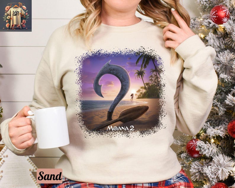 May include: A white sweatshirt with a graphic print of a teal blue ocean scene with a large teal blue hook, a sunset, and palm trees. The text "Moana 2" is printed below the hook.
