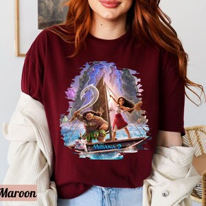 May include: A maroon t-shirt featuring a graphic print of Moana and Maui sailing on a boat. The graphic is surrounded by a blue and purple watercolour effect. The text "Moana 2" is printed on the boat.
