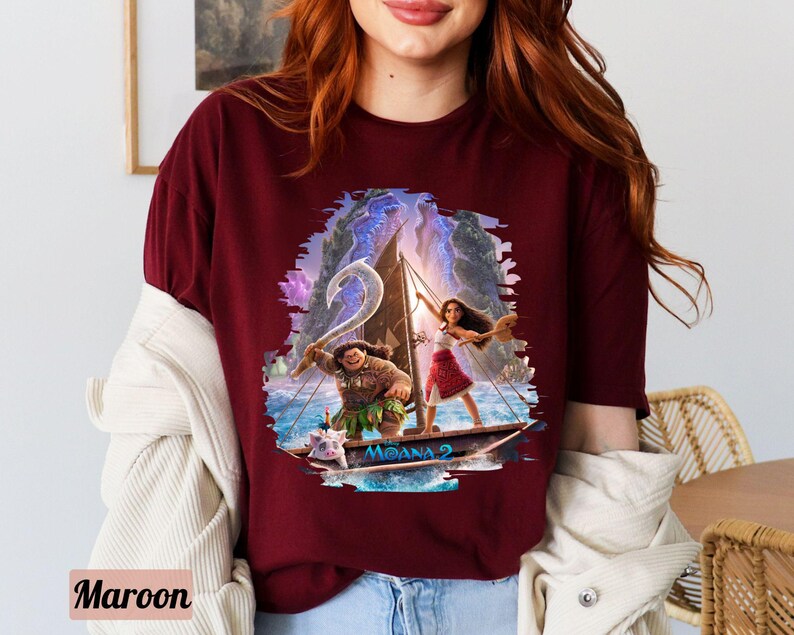 May include: A maroon t-shirt featuring a graphic print of Moana and Maui sailing on a boat. The graphic is surrounded by a blue and purple watercolour effect. The text "Moana 2" is printed on the boat.